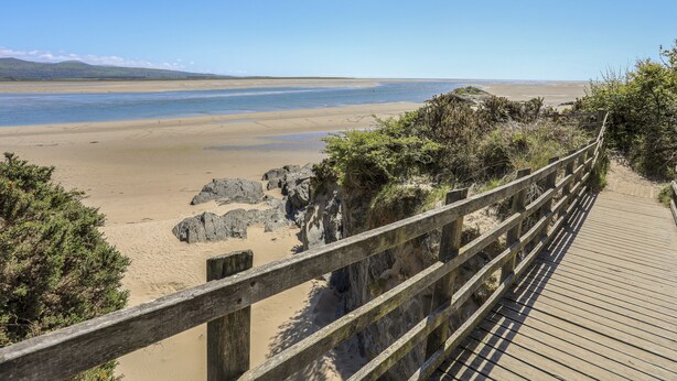 A wooden boardwalk leading to a sandy beach with rocky outcrops and sparse vegetation. - 31 Beach Road (Porthmadog)