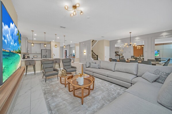 A spacious living room with a large sofa, a coffee table, and a TV on a stand. There is a kitchen area with a refrigerator and a dining table.