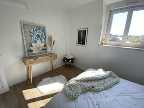 A bedroom with a bed, a bedside table with a vase, a framed painting, and a window with a view. - A spacious, elegant apartment in the heart of Epernay. Quiet top floor with a magnificent view. (Épernay)