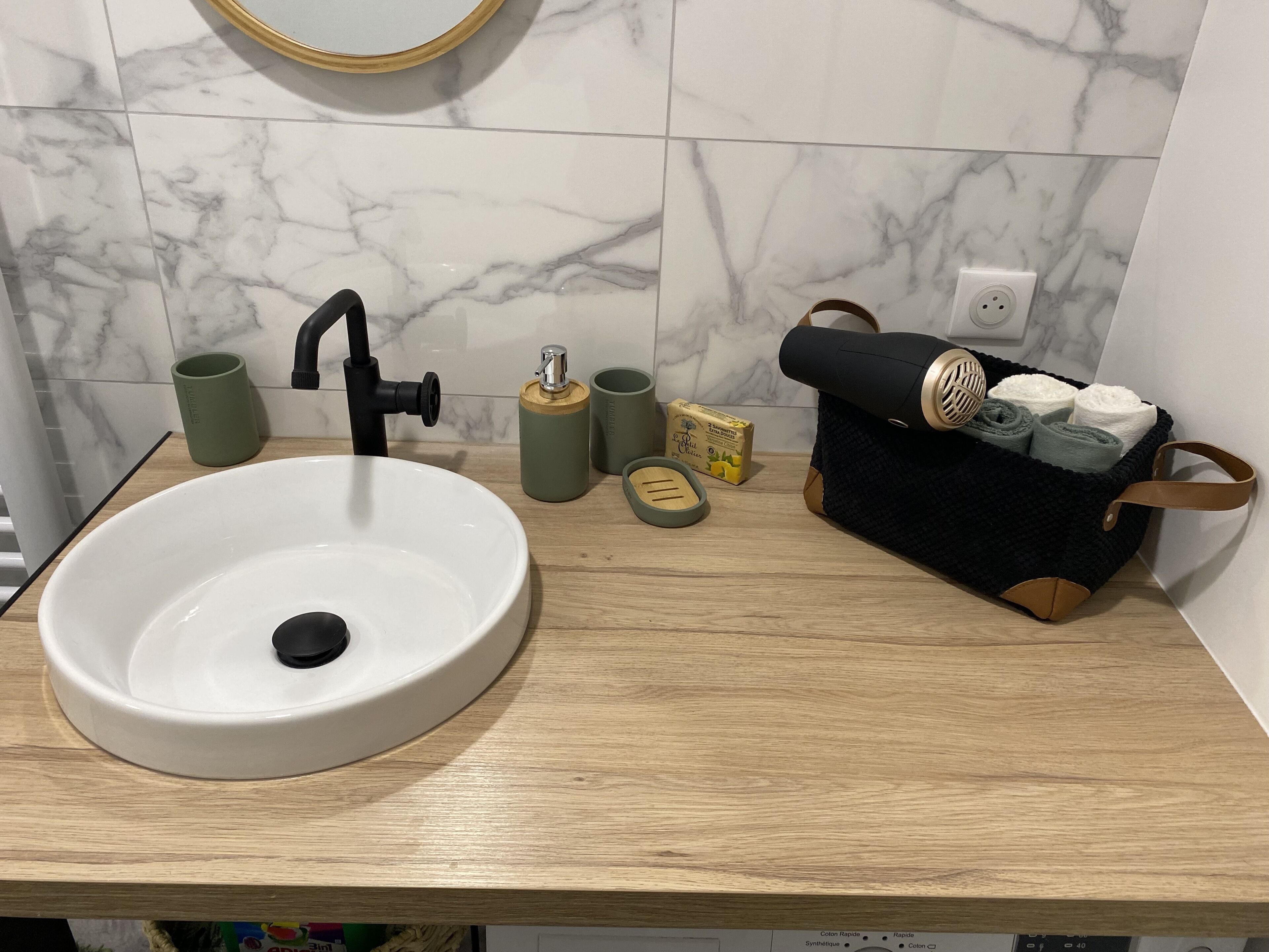 Ein Badezimmer mit einem weißen Waschbecken, einem schwarzen Wasserhahn, einer Holz-Arbeitsplatte, einem grünen Becher, einer hölzernen Seifenspender sowie einem schwarzen Handtuchhalter mit Handtüchern und einer gelben Seifenschale.