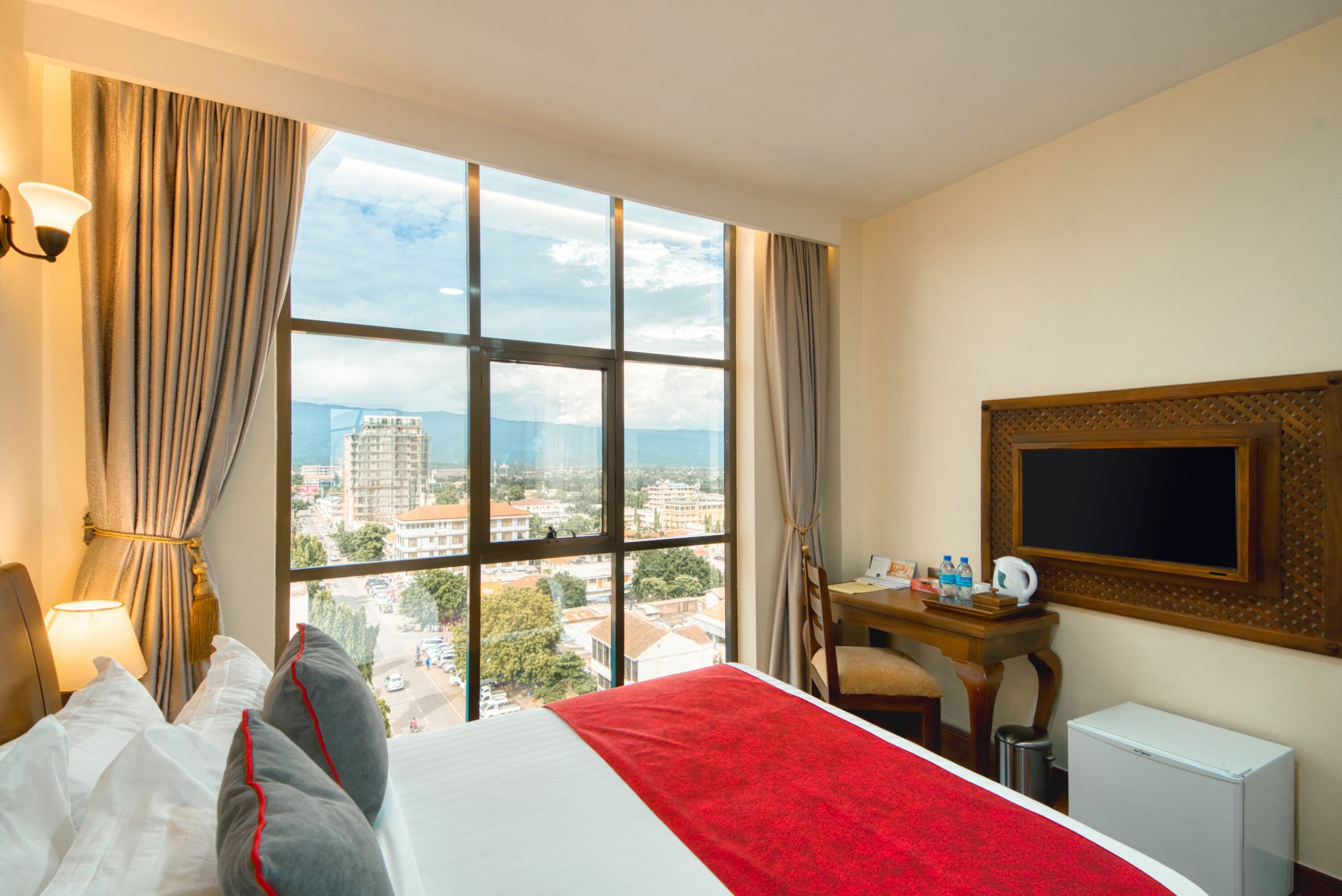 Quarto de hotel com uma janela grande, cama com roupa de cama branca e vermelha, escrivaninha com cadeira e radiador branco pequeno.
