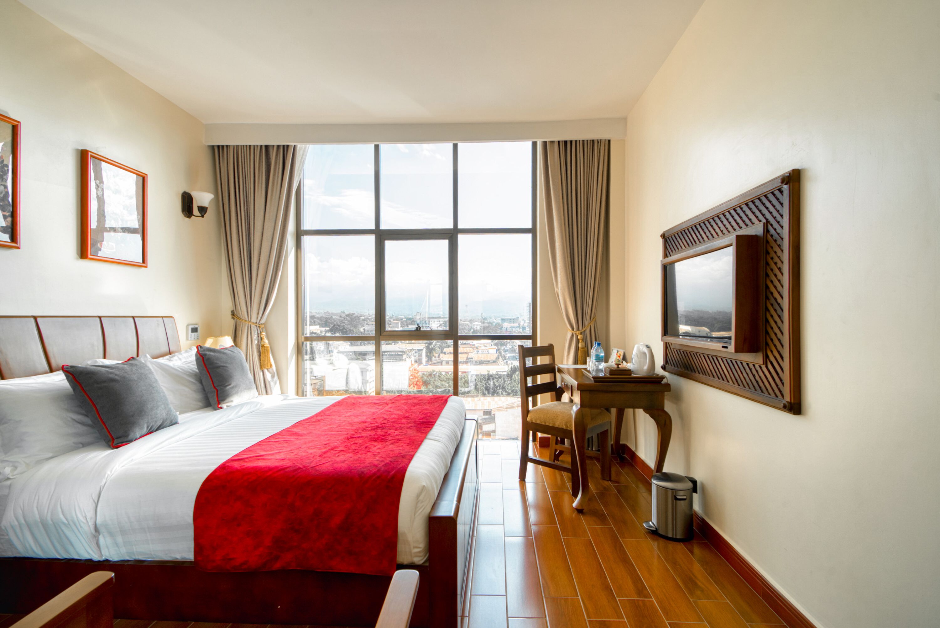 A hotel room with a large bed, a desk with a chair, a TV mounted on the wall, and a view of the city through large windows.