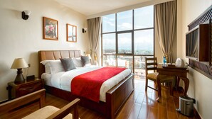 A hotel room with a large bed, a desk, a chair, a TV, and a balcony with a city view. - Kibo Palace Hotel Moshi (Moshi)