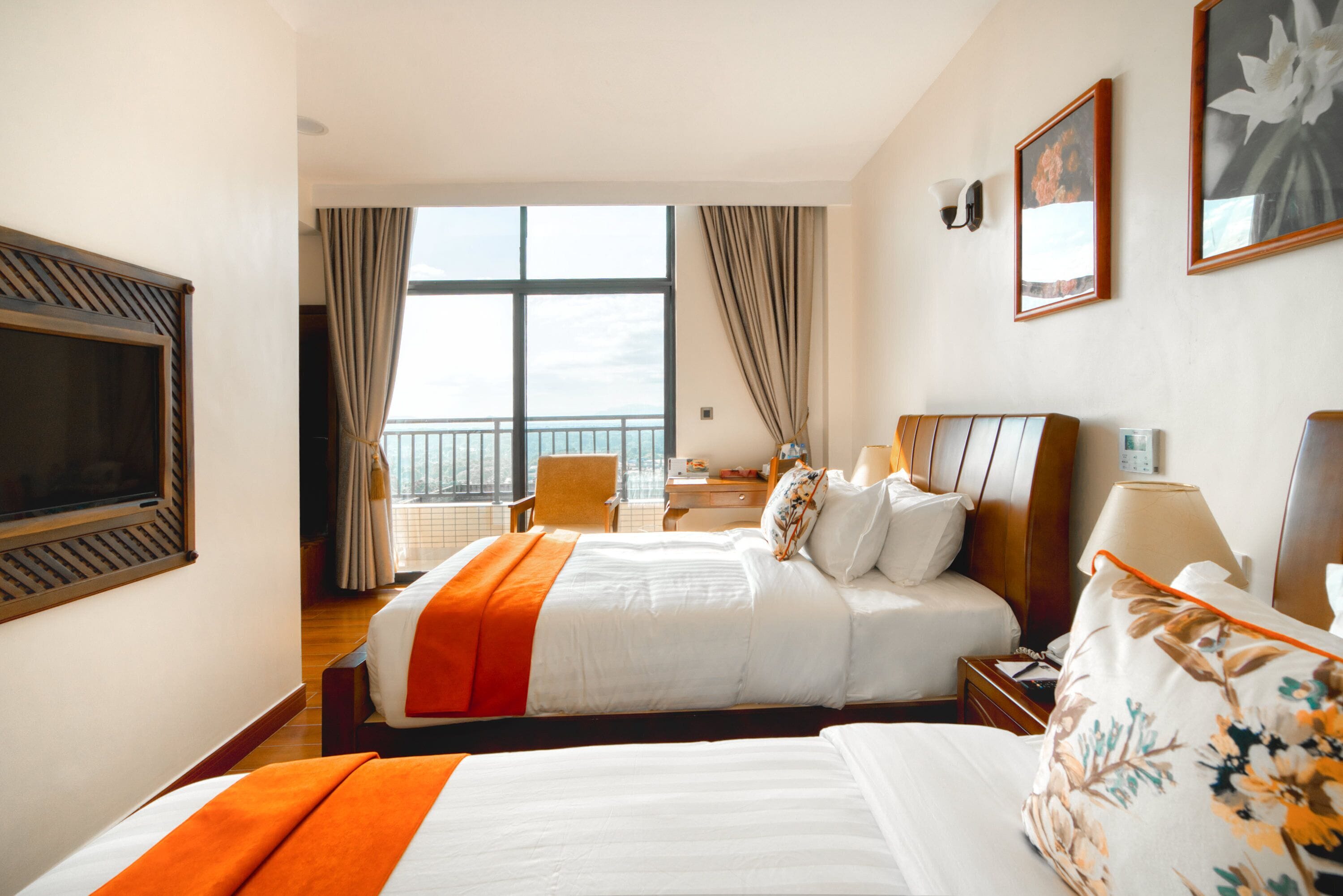 Habitación de hotel con dos camas, televisión, escritorio y balcón con cortinas.