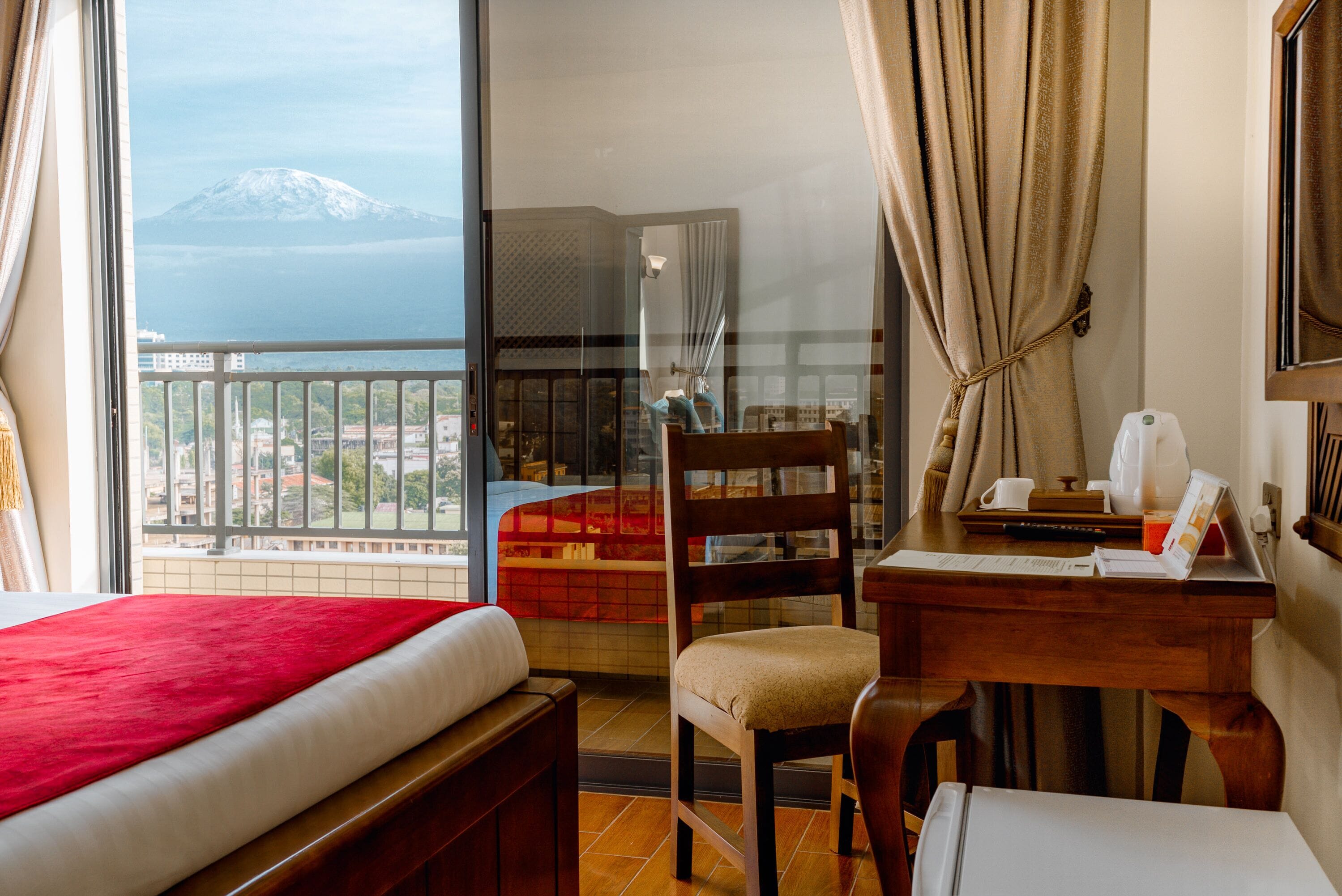 A hotel room with a bed, a desk with a chair, a window with a view of a mountain, and a balcony with a railing.