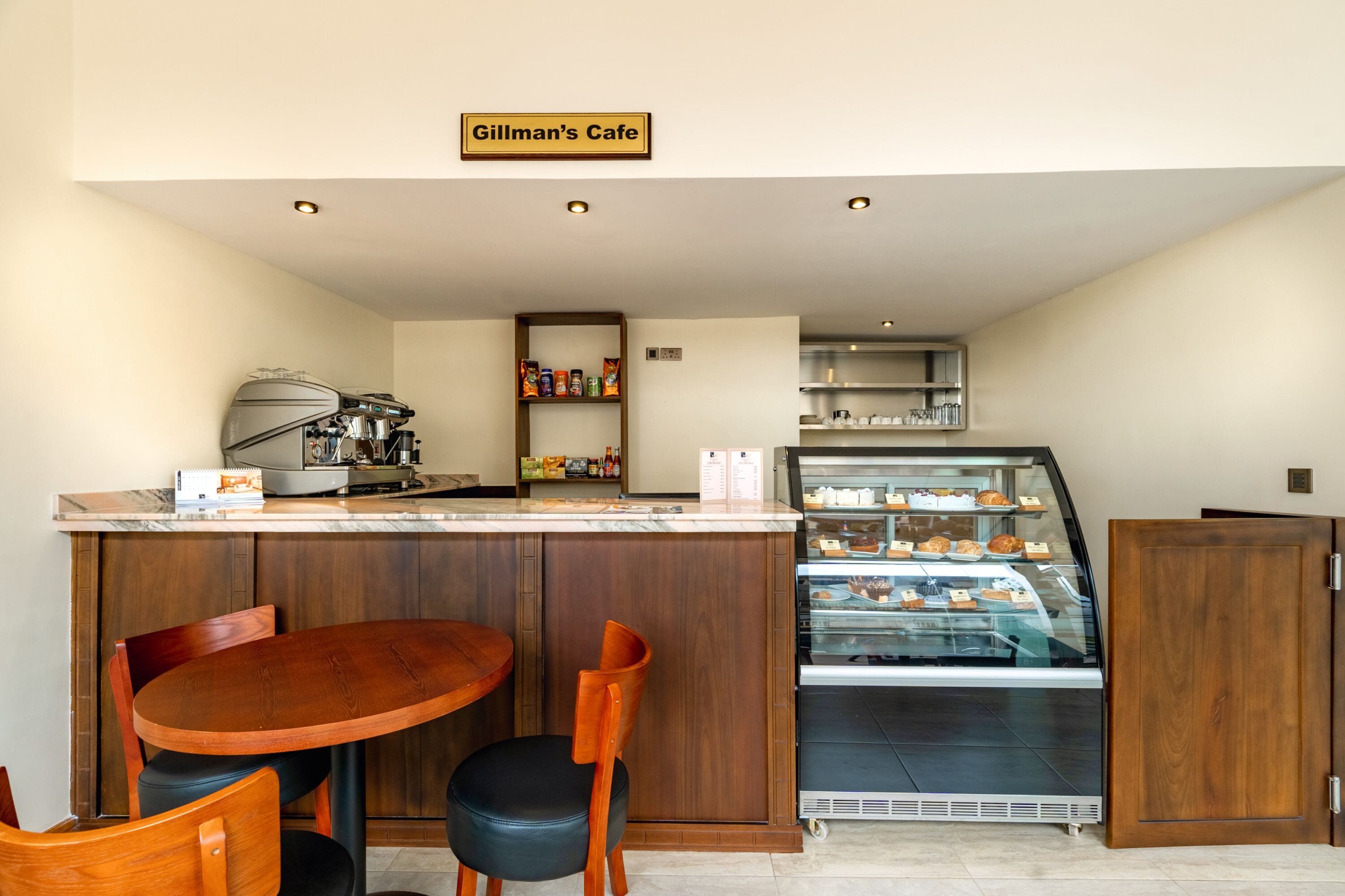 Un café avec une table et des chaises en bois, un appareil à café, une vitrine remplie de pâtisseries et une enseigne indiquant « Café Gillman ».