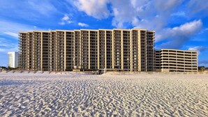 Ein mehrstöckiges Hotel mit flachem Dach und Balkonen, gelegen an einem Sandstrand unter blauem Himmel.