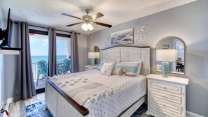 Ein Schlafzimmer mit Bett, Kommode, Deckenventilator, einem Fenster mit Vorhängen und Blick aufs Meer.