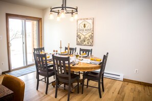 A dining room with a wooden table set for six, surrounded by dark chairs. A chandelier hangs above, and a framed picture of a bee with the text "FRESH HONEY" is on the wall.