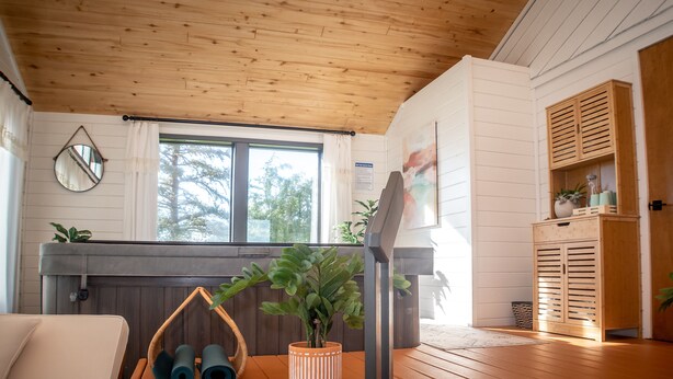 A cozy room with a wooden ceiling, a large window, a sofa, a small table with yoga mats, a potted plant, and a wooden cabinet.