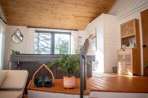 Una stanza accogliente con soffitto in legno, un'ampia finestra, un divano, un tavolino con tappetini da yoga, una pianta in vaso e un armadietto in legno.