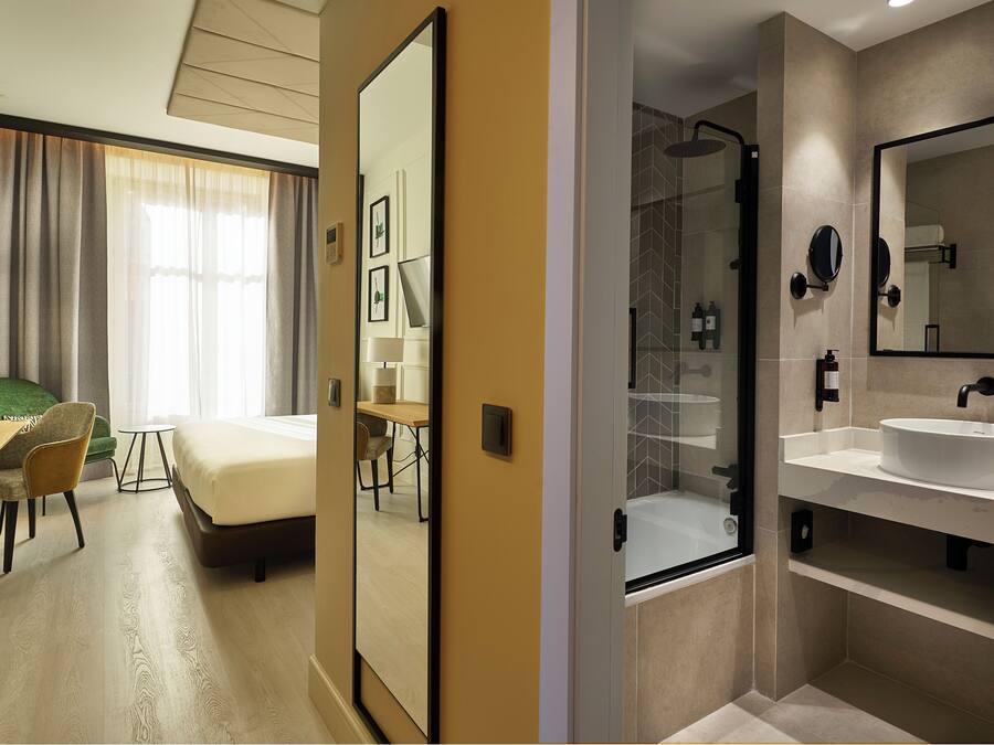 A hotel room with a bed, a desk, a chair, a dining table, and a bathroom with a sink and mirror.