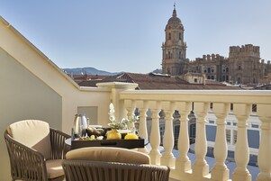 Un balcon avec un fauteuil en osier, un plateau avec de la nourriture et des boissons, et une vue sur des bâtiments d’architecture historique.