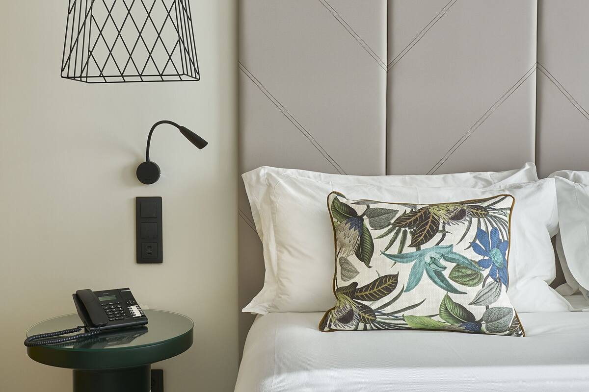 A modern bedroom with a bed, a decorative pillow, a wall-mounted light fixture, a telephone, and a small side table.