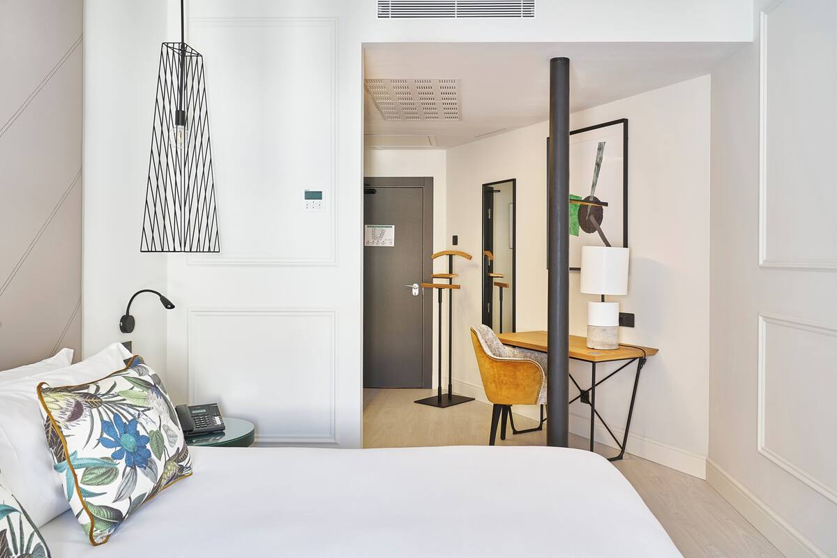 A hotel room with a bed, a desk, a chair, a lamp, and a hanging light fixture.
