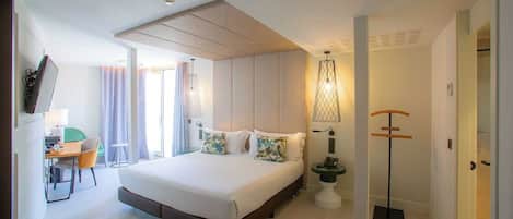 A modern hotel room with a large bed, a desk, and a chair. There is a lamp and a coat rack.