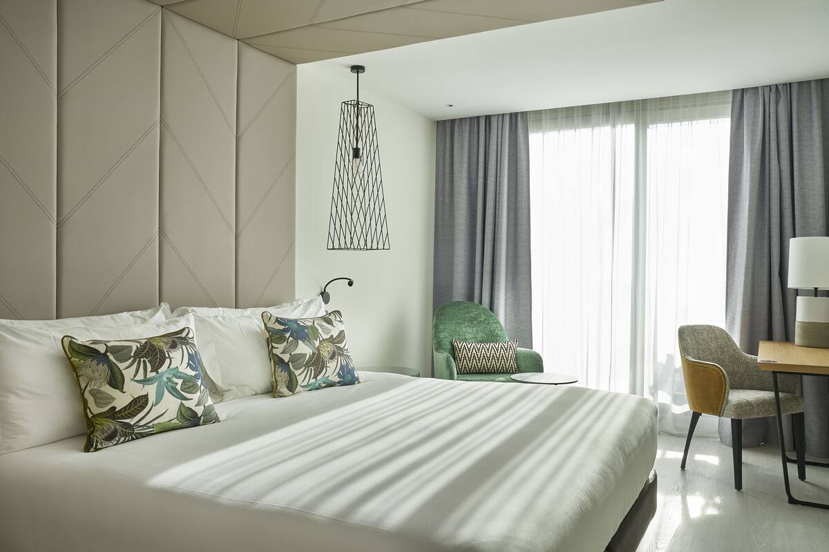 A modern hotel room with a large bed, a green armchair, a wooden desk, and a view of the outdoors through large windows.