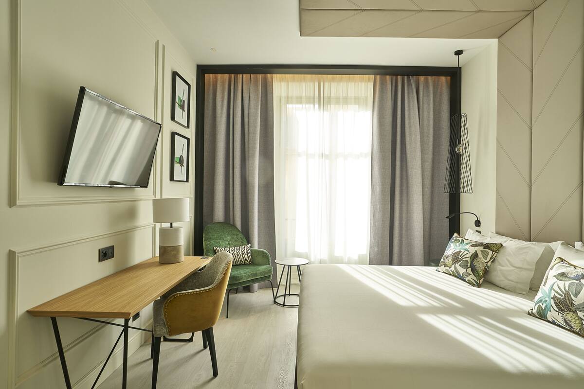 A hotel room with a bed, a desk, a chair, a TV mounted on the wall, and a window with curtains.