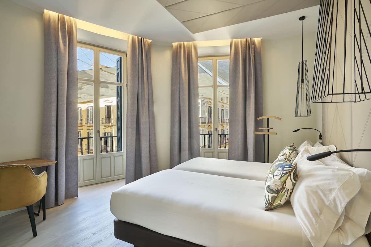 A hotel room with a large bed, two bedside tables, a chair, and a view of the city through large windows with curtains.