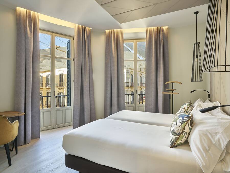 A hotel room with a large bed, two bedside tables, a chair, and a view of the city through large windows with curtains.
