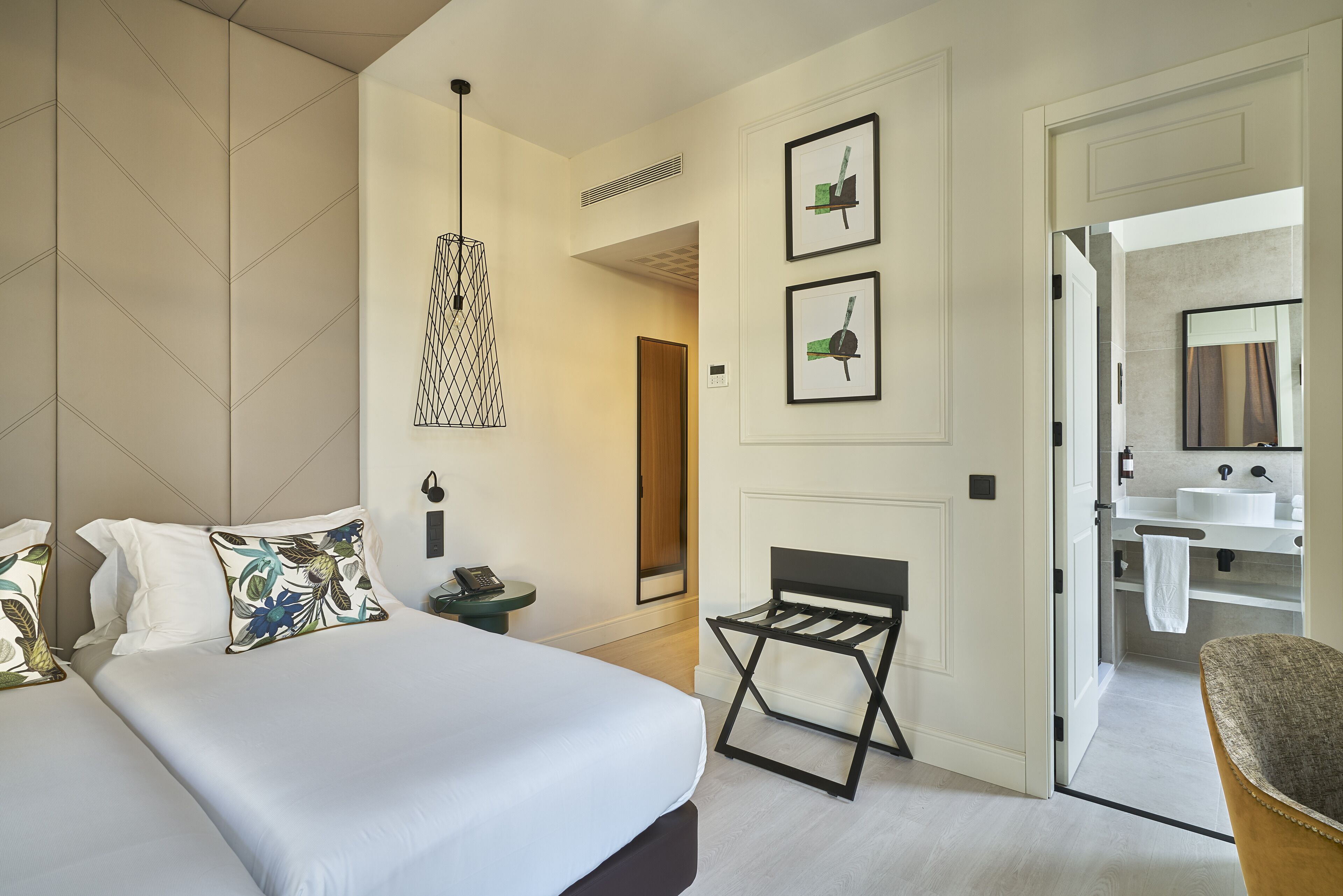 A hotel room with a bed, a small table, a chair, a hanging light fixture, and a bathroom visible through an open door.