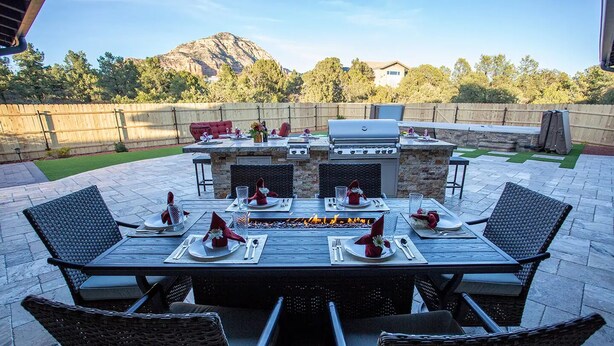 Un patio al aire libre con una mesa de piedra para cuatro personas, rodeado de sillas de mimbre, una parrilla y una chimenea de piedra.