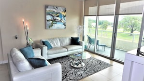 Un salón moderno con un sofá blanco, una mesa de centro de vidrio y un ventanal que ofrece vistas a un balcón con mobiliario exterior.