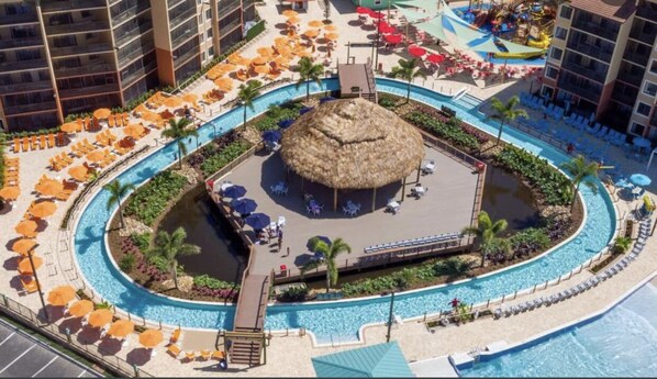 Relax by the central pool surrounded by lounge chairs and umbrellas, with a shaded area under a thatched-roof structure.