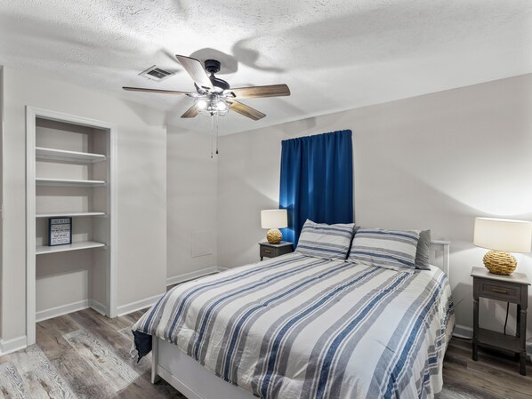Quarto com cama, ventoinha no teto, mesa de cabeceira, luminária, estante e cortina azul.
