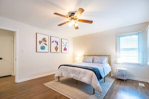 Un dormitorio con cama, ventilador de techo, ventana con persianas y tres cuadros en la pared.