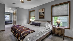 A bedroom with a large bed, a nightstand with a lamp, a ceiling fan, and a window with blinds. - Charming Modern Victorian in the Village of EVL (Ellicottville)