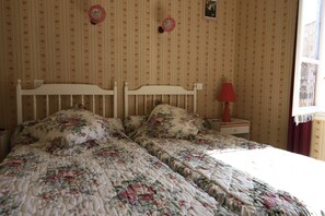 Quarto com papel de parede estampado com flores, uma cama branca com roupa de cama florida, uma mesa de cabeceira com abajur e uma janela com cortinas.