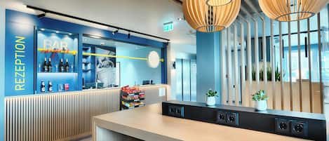 A hotel reception area with a blue wall, a reception desk, and a bar. There are plants and a blue chair in the foreground.