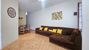 A living room with a brown sofa, yellow cushions, a dining table with chairs, a hanging light fixture, and a wall with a floral painting. - Piscina Aquecida, Ar Condicionado, Área Gourmet, Bilhar (Itanhaém)