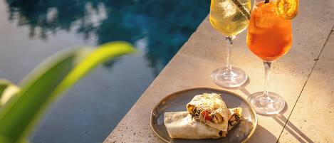 Une table dressée au bord de la piscine, avec une assiette de nourriture, deux boissons et une vue dégagée sur la piscine.