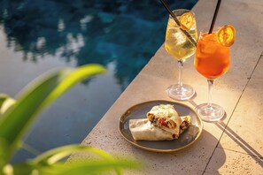 Escena junto a la piscina con un plato de comida, dos bebidas y vista despejada a la piscina.