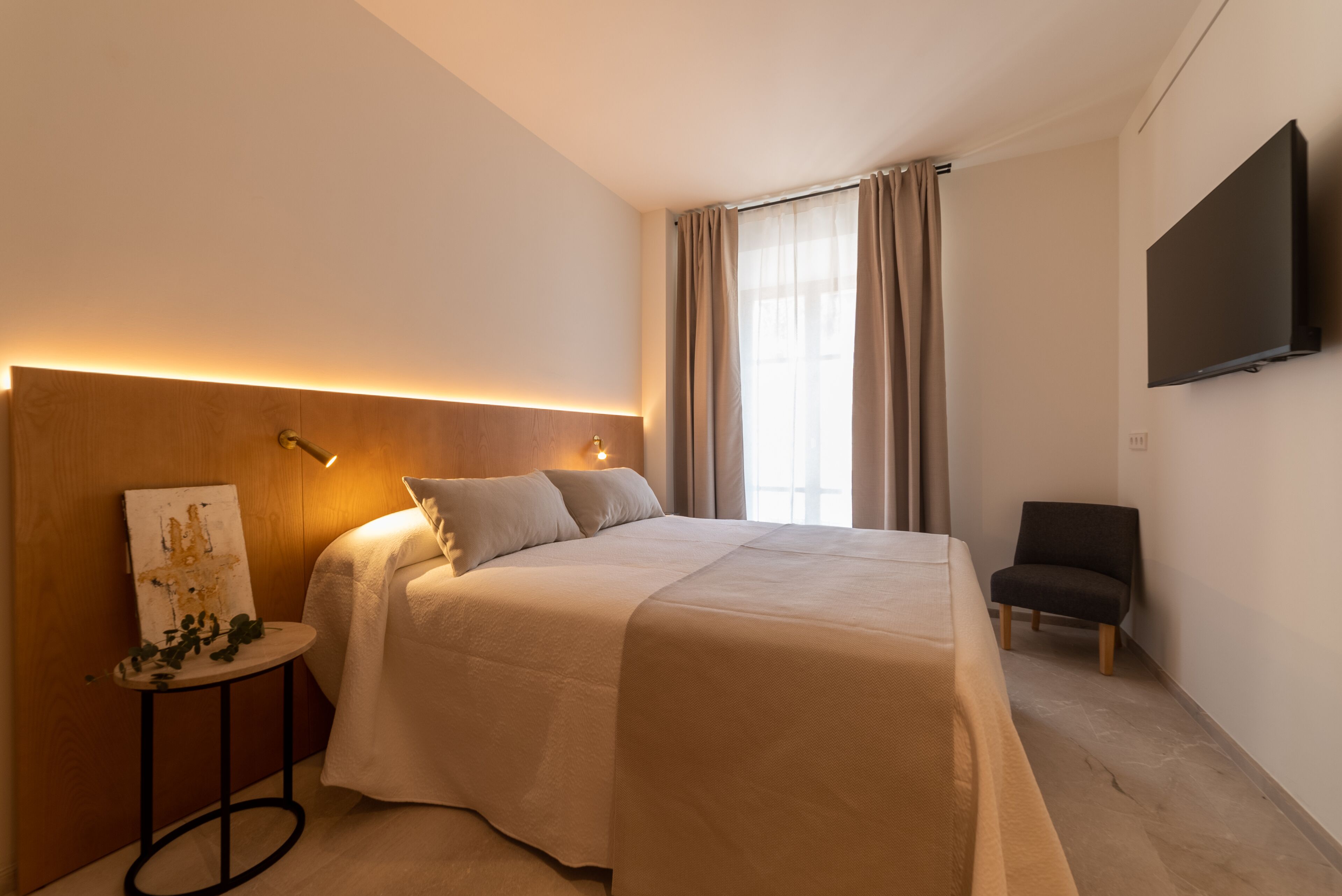 Ein modernes Hotelzimmer mit einem großen Bett, einem kleinen Beistelltisch mit einem Gemälde, einem Sessel und einem an der Wand montierten Flachbildfernseher.