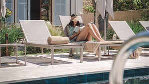 Un área junto a la piscina con sillones de descanso, una persona leyendo y un edificio con contraventanas azules al fondo.