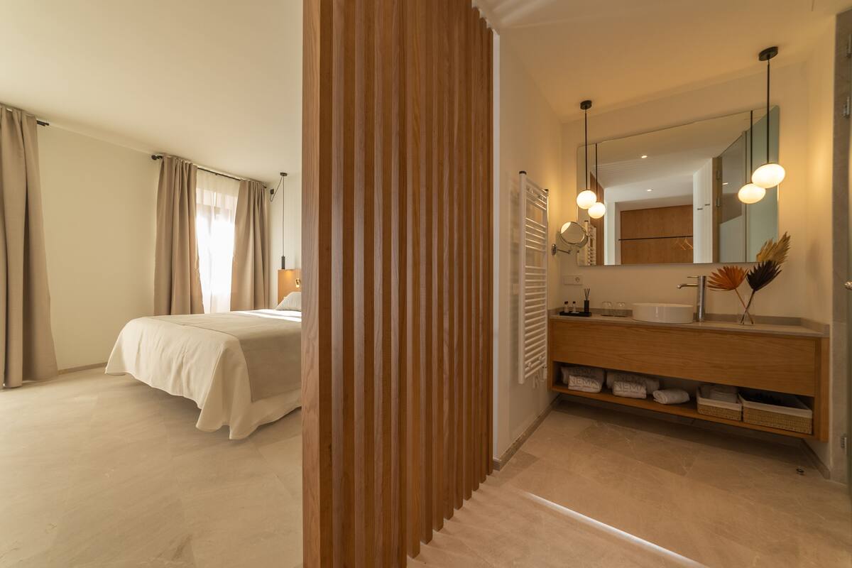Een moderne hotelkamer met een groot bed, een houten scheidingswand, een badkamer met een grote spiegel en een wastafel met opbergruimte.