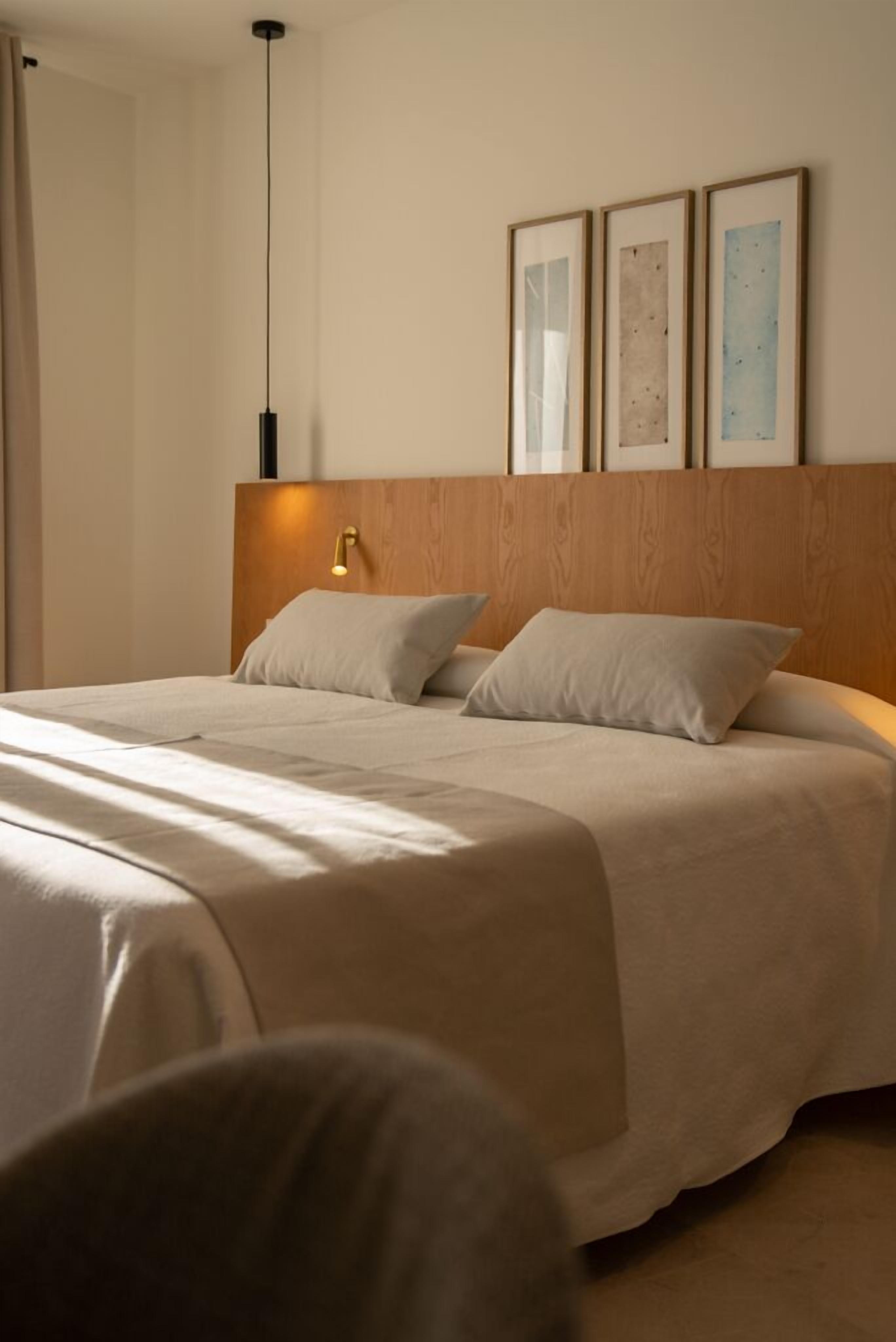 Una cama bien hecha con cabecera de madera, una lámpara de noche y tres cuadros en la pared.