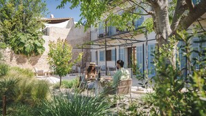 Un café al aire libre con mesas y sillas, rodeado de vegetación y un edificio con contraventanas azules.