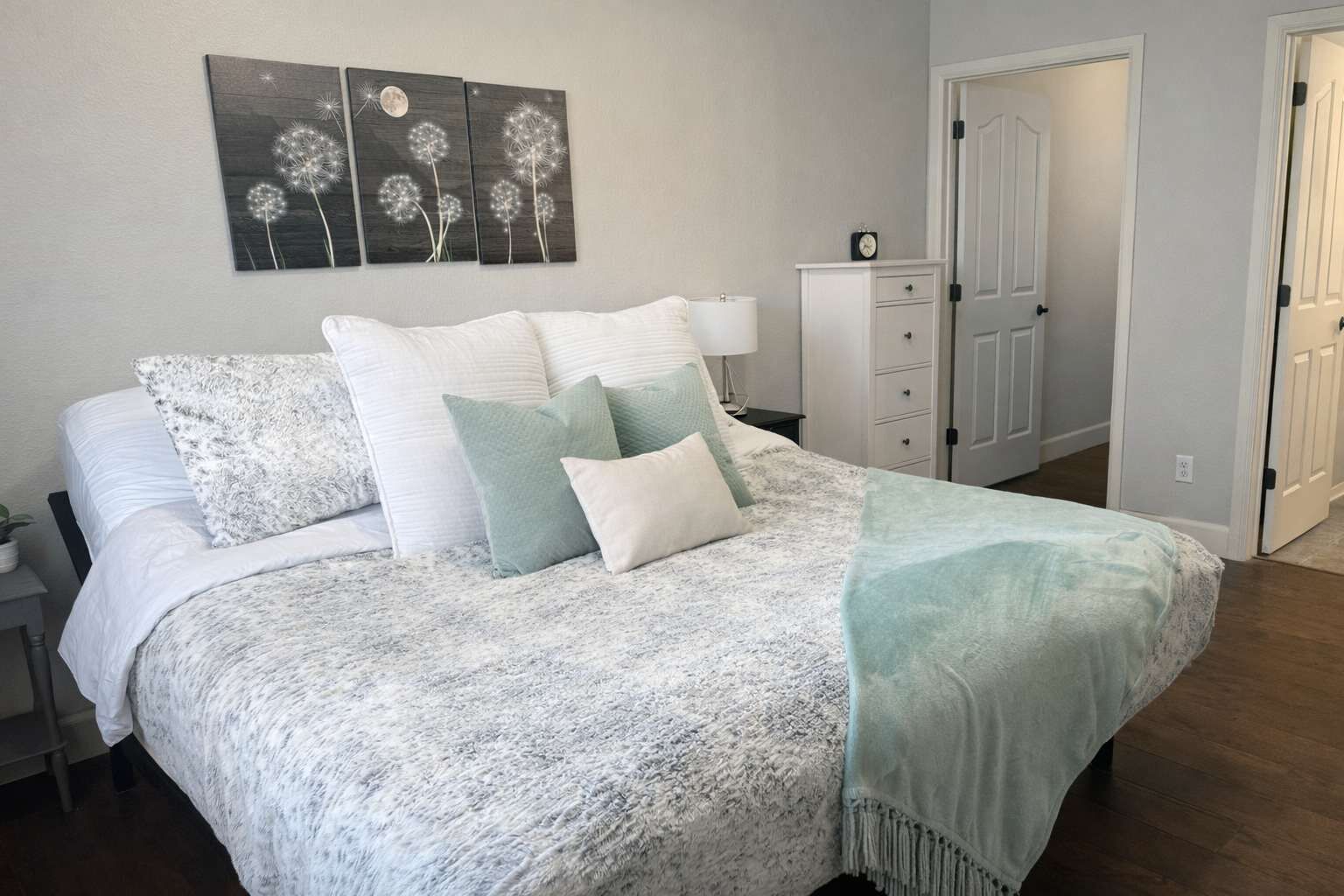 A neatly made bed with a patterned bedspread and pillows, a bedside table with a lamp, and a wall-mounted artwork of dandelions.