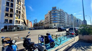 Una calle con edificios, un autobús y vehículos estacionados, incluyendo una patineta y una bicicleta.