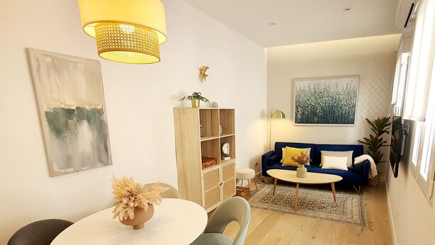 Una sala de estar moderna con una mesa de comedor redonda, dos sillas, un sofá azul con cojines, una estantería y un cuadro abstracto grande en la pared.