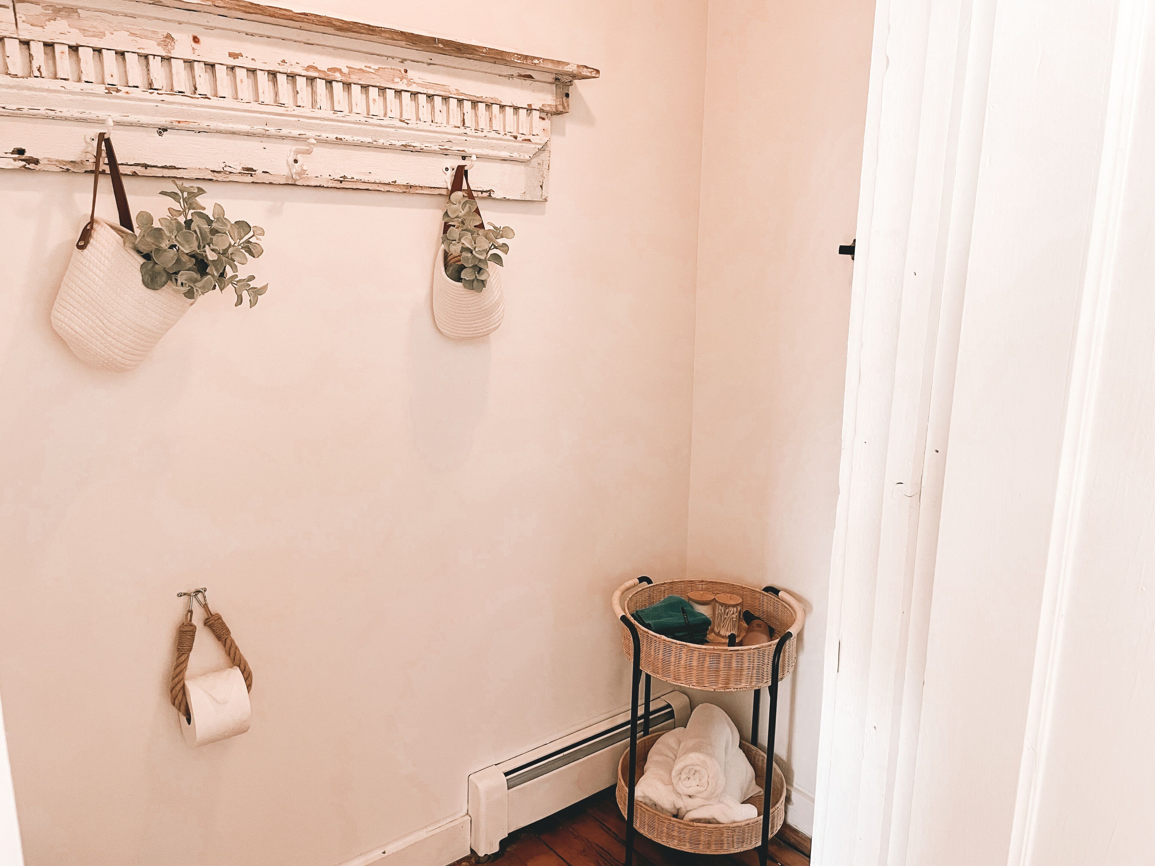 Un baño con un radiador blanco, un estante de mimbre con toallas, y dos cestas colgantes con plantas.