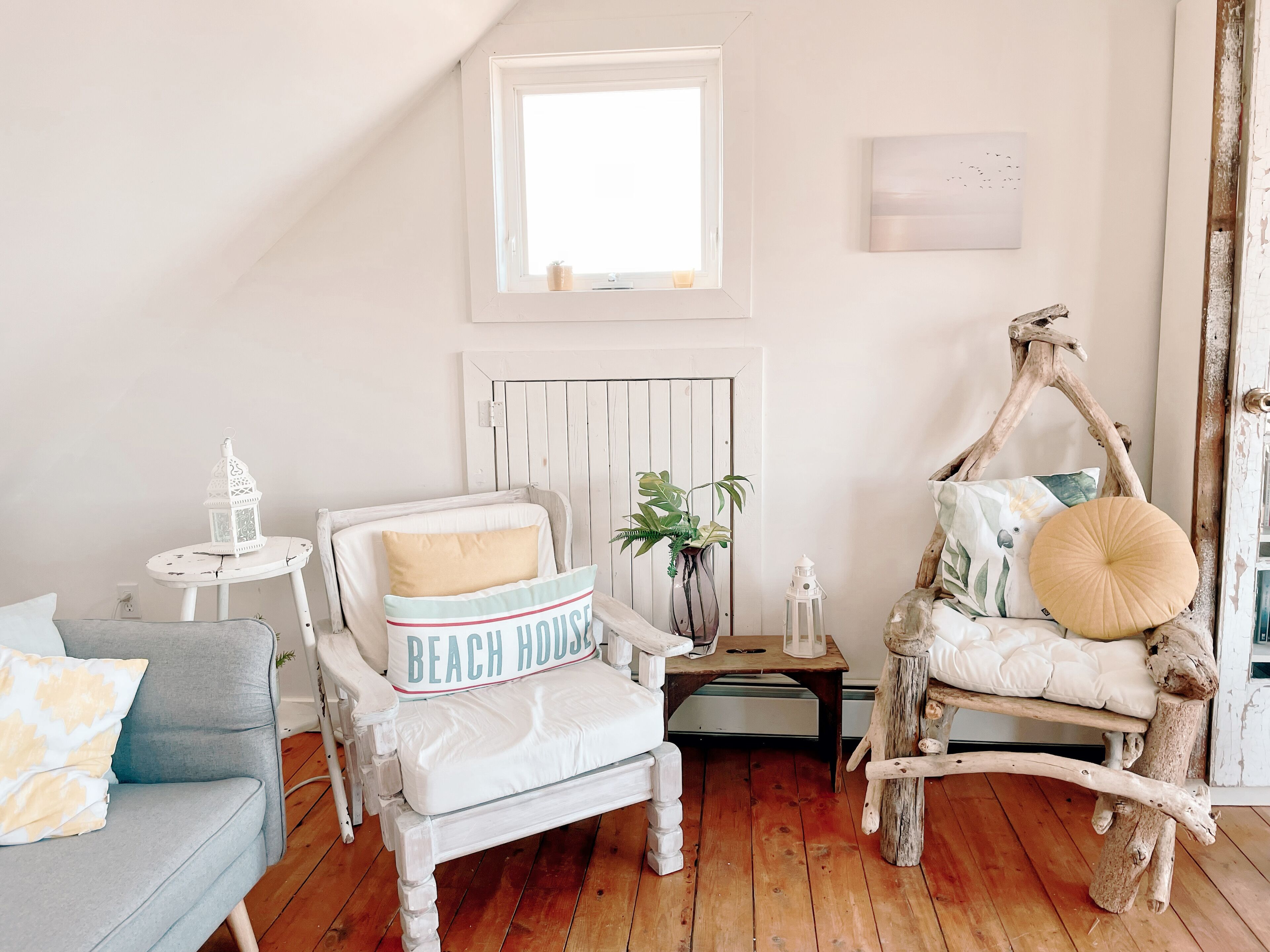 A cozy room with a white chair, a small table, and a rustic wooden chair. A window provides natural light, and a radiator is mounted on the wall.