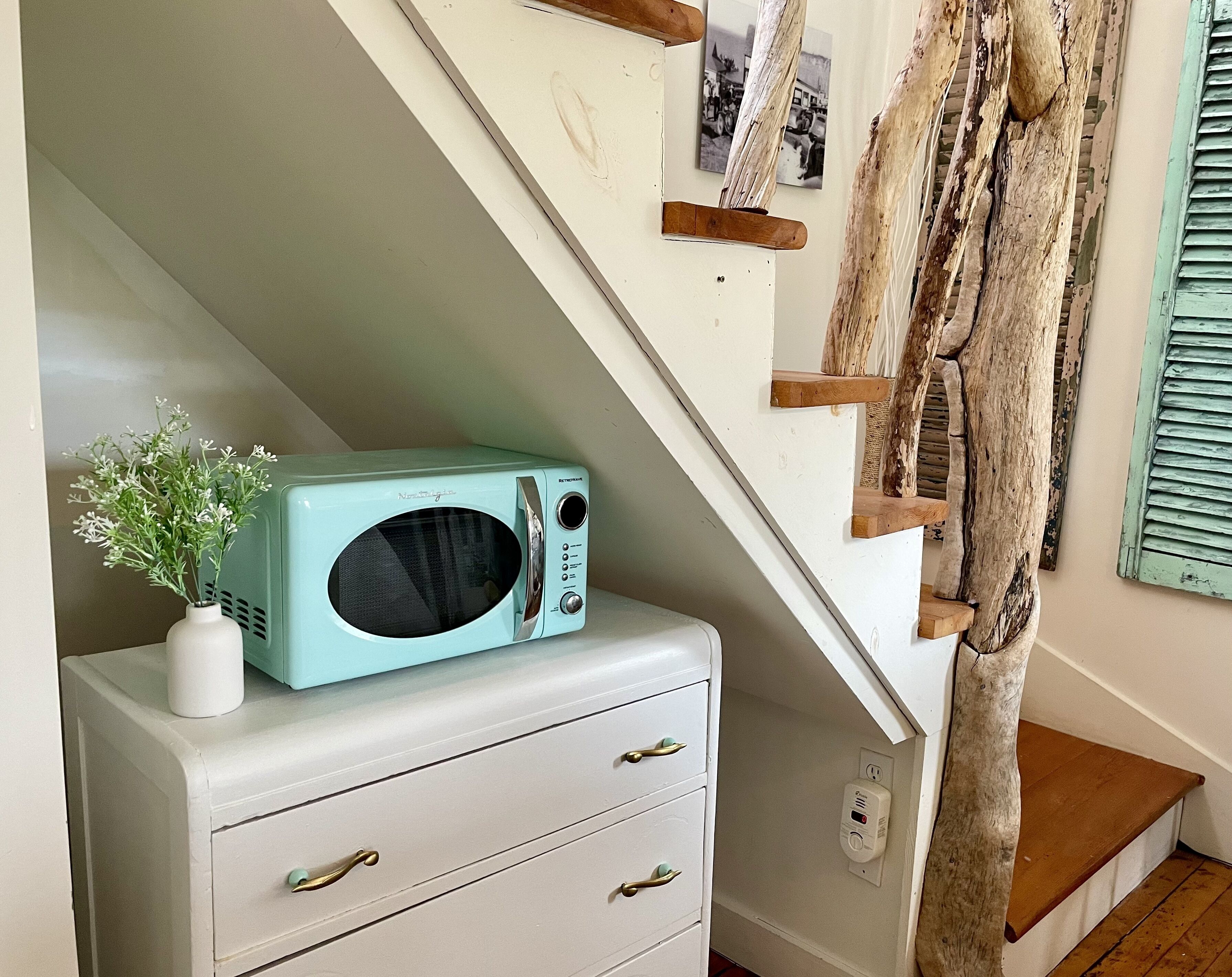 A teal-colored microwave on a white dresser, a vase with flowers, and a rustic wooden staircase.