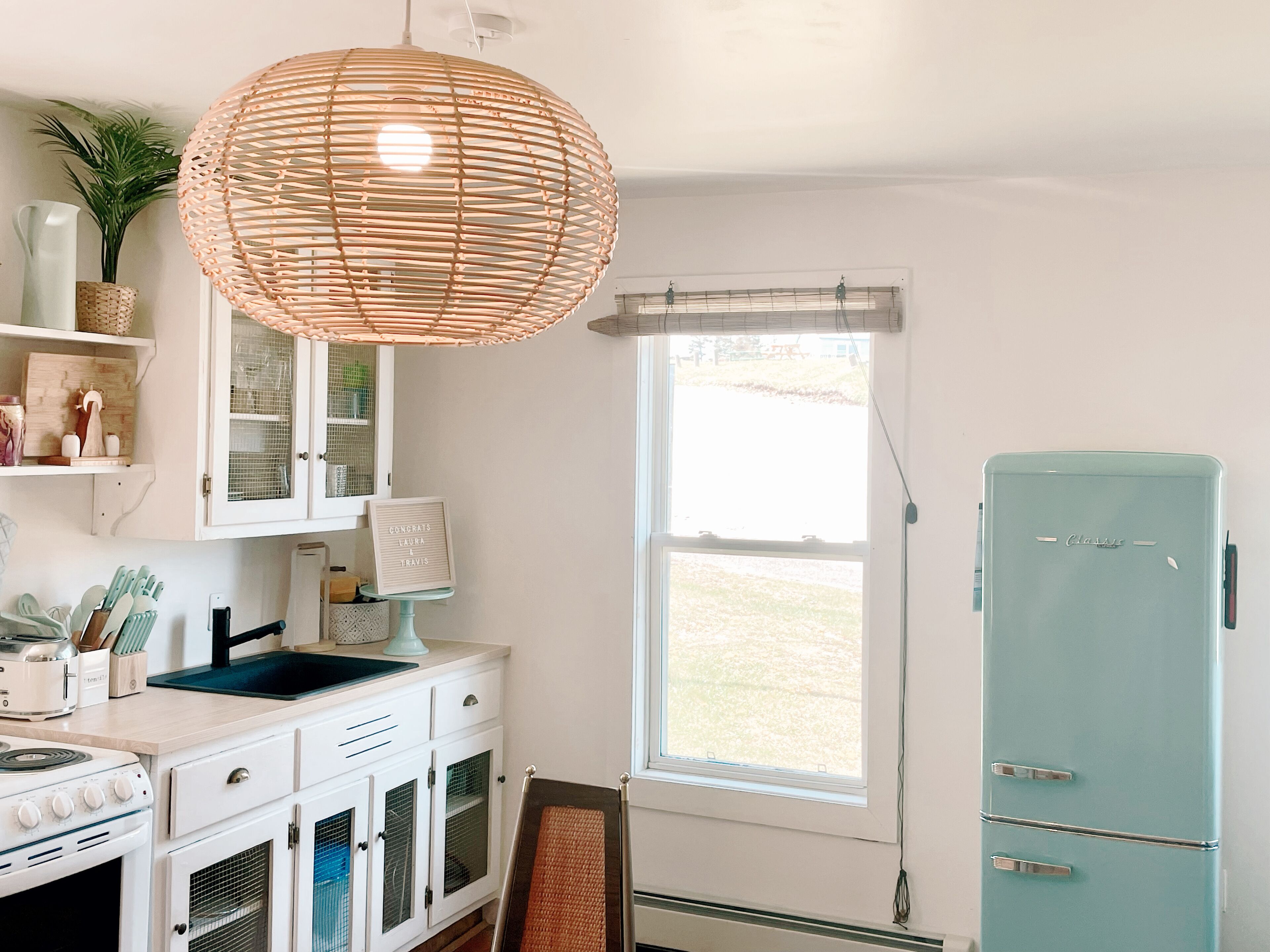 A kitchen with white cabinets, a blue refrigerator, a hanging light fixture, and a window with blinds.