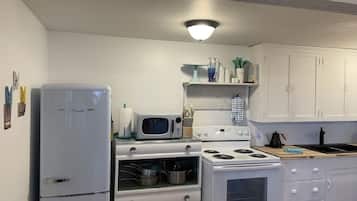 Uma cozinha com eletrodomésticos brancos, incluindo frigorífico, micro-ondas e fogão. Há armários e uma prateleira com diversos utensílios de cozinha.
