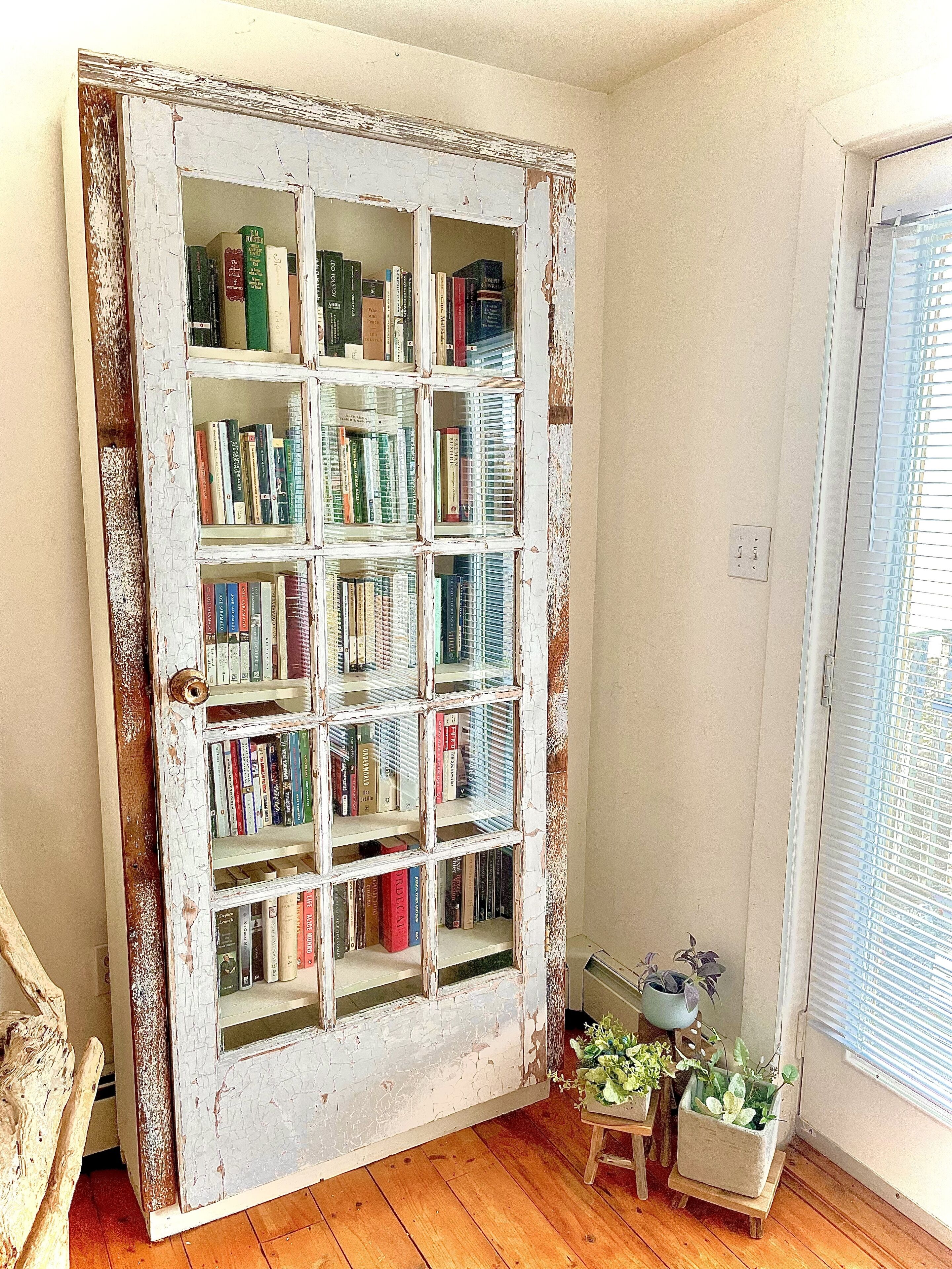 A rustic bookshelf with multiple shelves filled with books, a small wooden stool with potted plants, and a window with blinds.